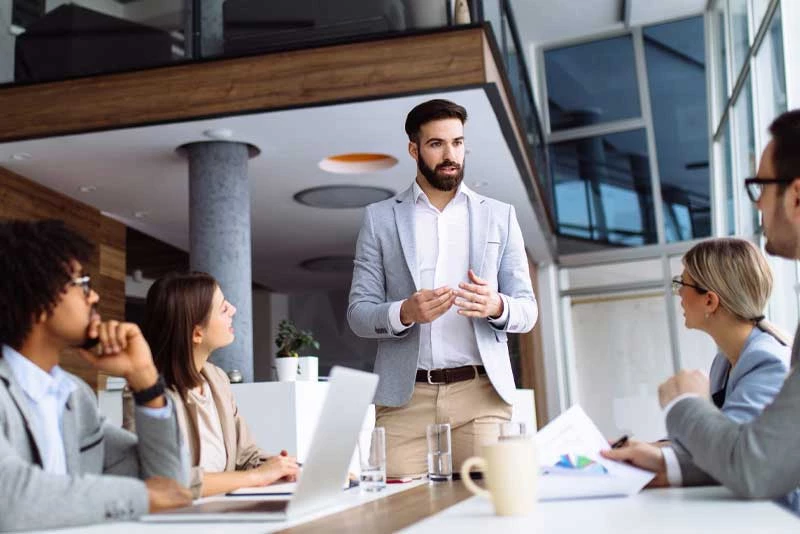A group of colleagues with laptops and charts meet around a table to discuss syndicated loans
